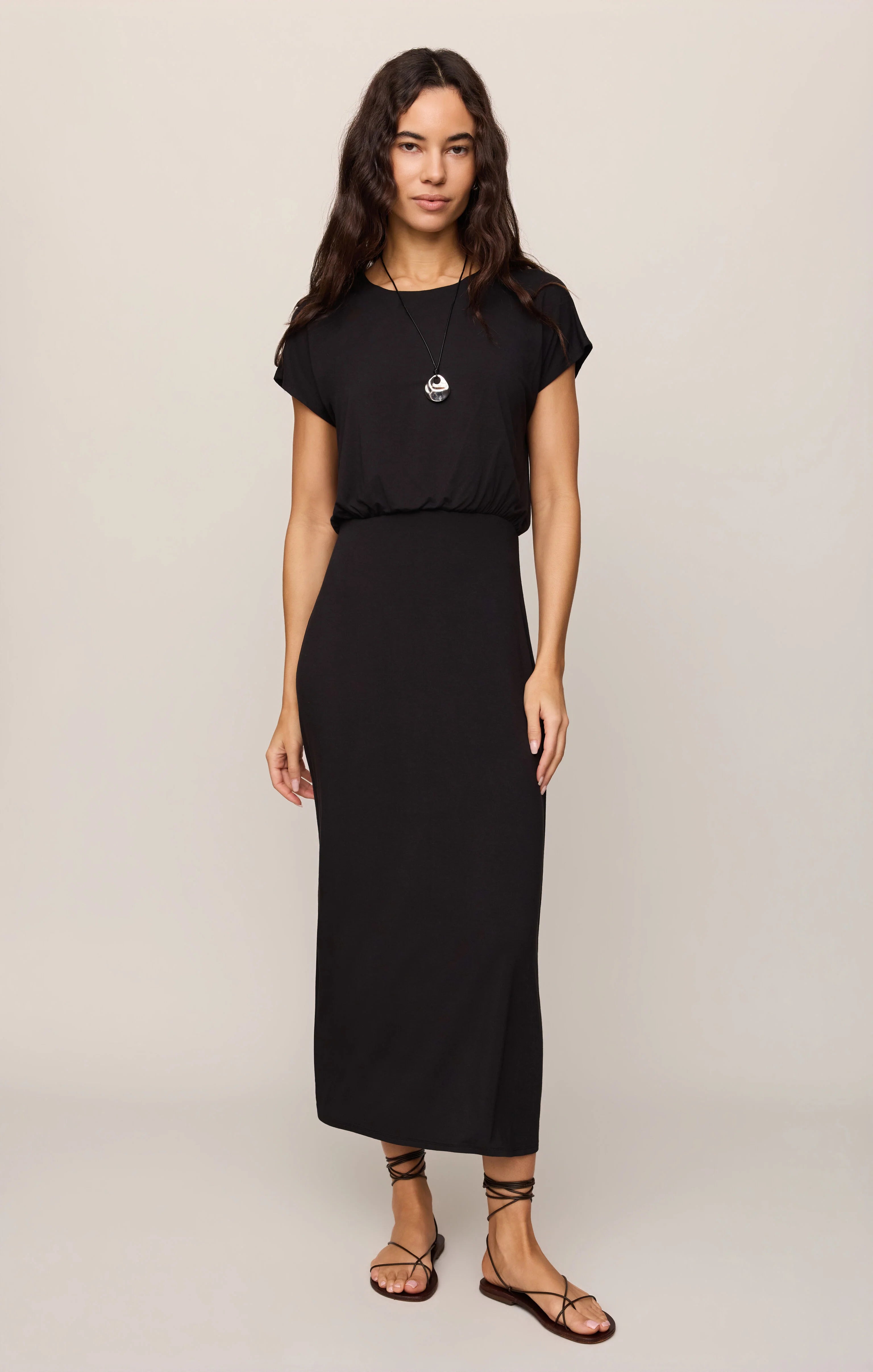 A woman modeling a black midi dress with short sleeves and a gathered waist, paired with brown strappy flat sandals against a light beige studio background.