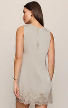 A woman shown from the back wearing a sleeveless light taupe-gray mini dress with a lace hem, photographed against a plain beige studio background.