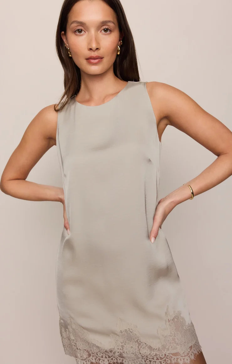 A woman modeling a sleeveless taupe-gray dress with a subtle sheen and a lace hem, set against a solid light beige background.