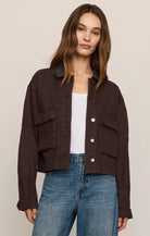 A woman modeling a dark brown cropped utility jacket with front flap pockets, worn over a white top and blue jeans, against a plain beige studio background.