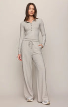 A woman modeling a light gray two-piece loungewear set consisting of a long-sleeve henley top and matching wide-leg sweatpants, photographed against a neutral light gray studio background.