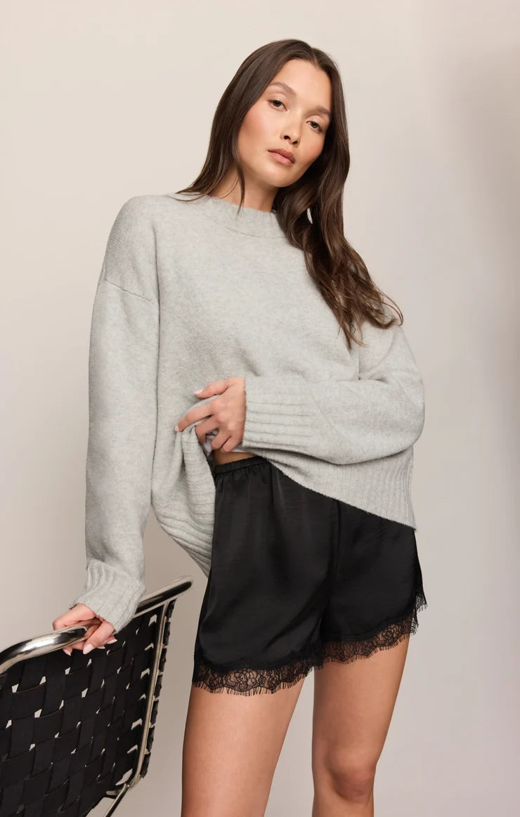 A woman modeling a light gray oversized knit sweater and black satin shorts with lace trim, standing against a plain light beige studio background and holding a black studded chair.