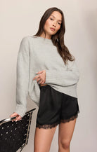 A woman modeling a light gray oversized knit sweater and black satin shorts with lace trim, standing against a plain light beige studio background and holding a black studded chair.