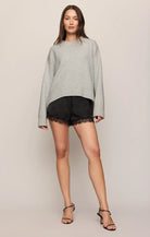 A woman modeling a light gray oversized knit sweater and black lace-trimmed shorts against a plain beige studio background.