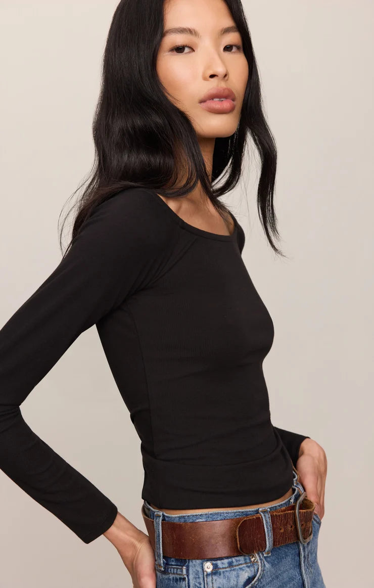 A woman modeling a fitted black long-sleeve scoop-neck top paired with blue jeans and a brown belt against a plain light beige studio background.