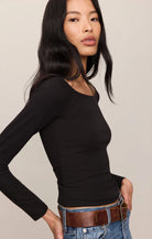 A woman modeling a fitted black long-sleeve scoop-neck top paired with blue jeans and a brown belt against a plain light beige studio background.
