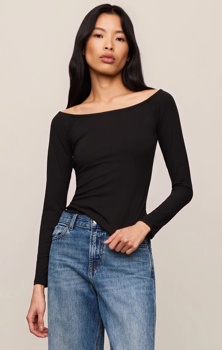 A woman modeling a black long-sleeve off-the-shoulder top paired with blue jeans, photographed against a plain light beige studio background.