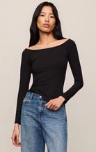 A woman modeling a black long-sleeve off-the-shoulder top paired with blue jeans, photographed against a plain light beige studio background.