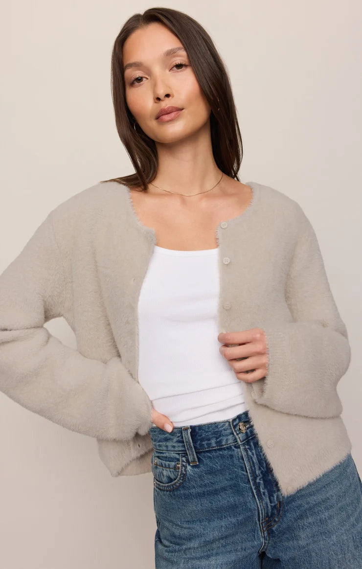 A woman wearing a light beige fuzzy knit cardigan over a white tank top and blue jeans, photographed against a plain light beige studio background.