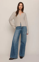 A woman modeling a light beige fuzzy knit cardigan with long sleeves and blue wide-leg jeans, standing against a plain light beige studio background.