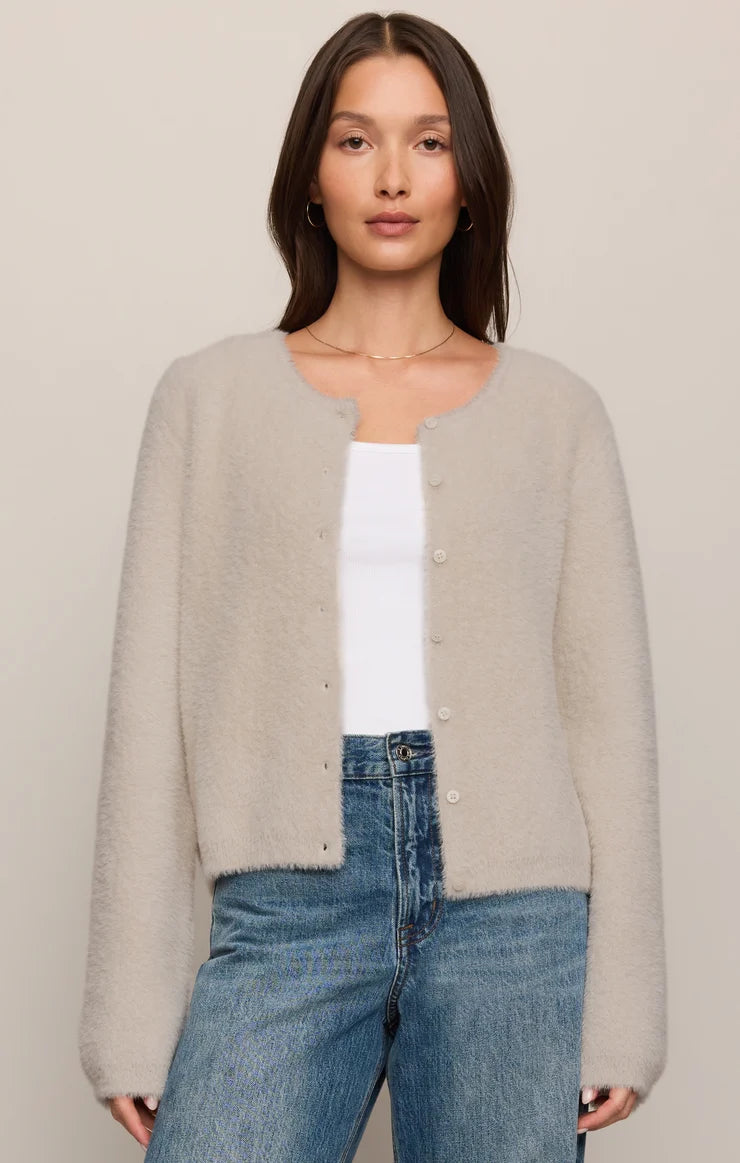 A woman modeling a light beige fuzzy knit cardigan with a round neckline and button-front closure, worn over a white top and blue jeans against a plain beige studio background.