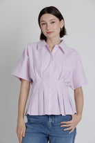 Woman wearing a light purple/pink button-up shirt and blue jeans against a plain background