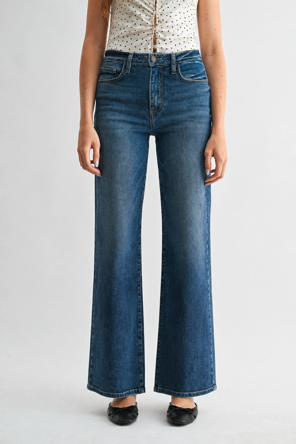 A model wearing a pair of high-waisted, wide-leg blue denim jeans with a faded wash, paired with a white polka-dot top and black flats, shown against a light gray studio background.