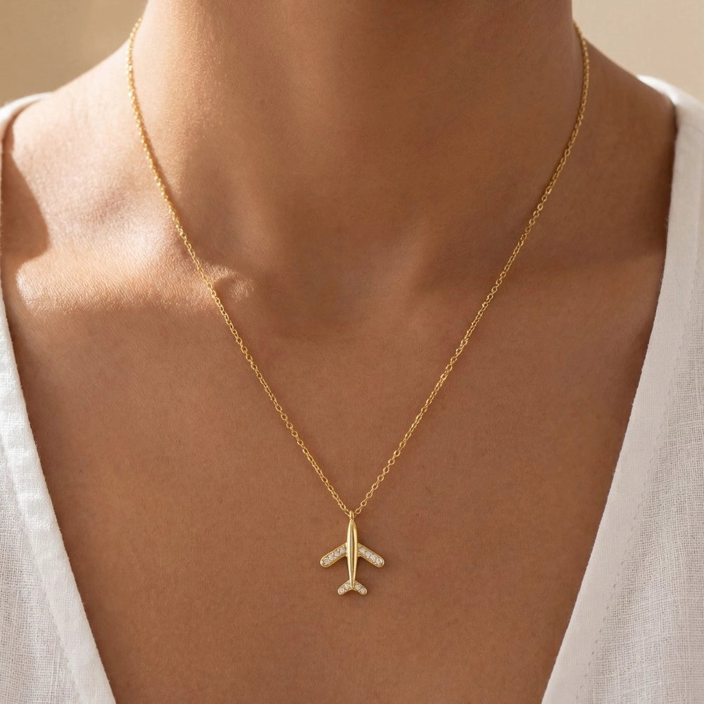 Gold airplane necklace on a person wearing a white top against a beige background