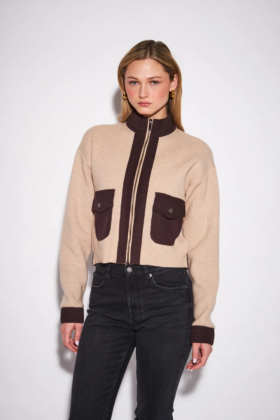 A woman modeling a cropped beige knit zip-up jacket with dark brown contrast panels, including the front zipper, pocket flaps, and cuffs, paired with black jeans against a plain light gray background.