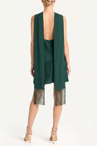 A model shown from the back wearing a dark green sleeveless open-front dress with fringe trim at the hem, paired with nude high-heeled sandals against a plain white studio background.