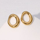 A pair of gold-toned, organic-shaped hoop earrings displayed on a white textured surface.