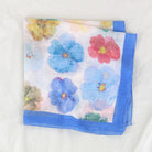 A square white scarf with a colorful watercolor floral print and a solid blue border, folded on a textured white fabric background.