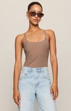 Woman wearing a brown bodysuit and light blue jeans with sunglasses on a beige background