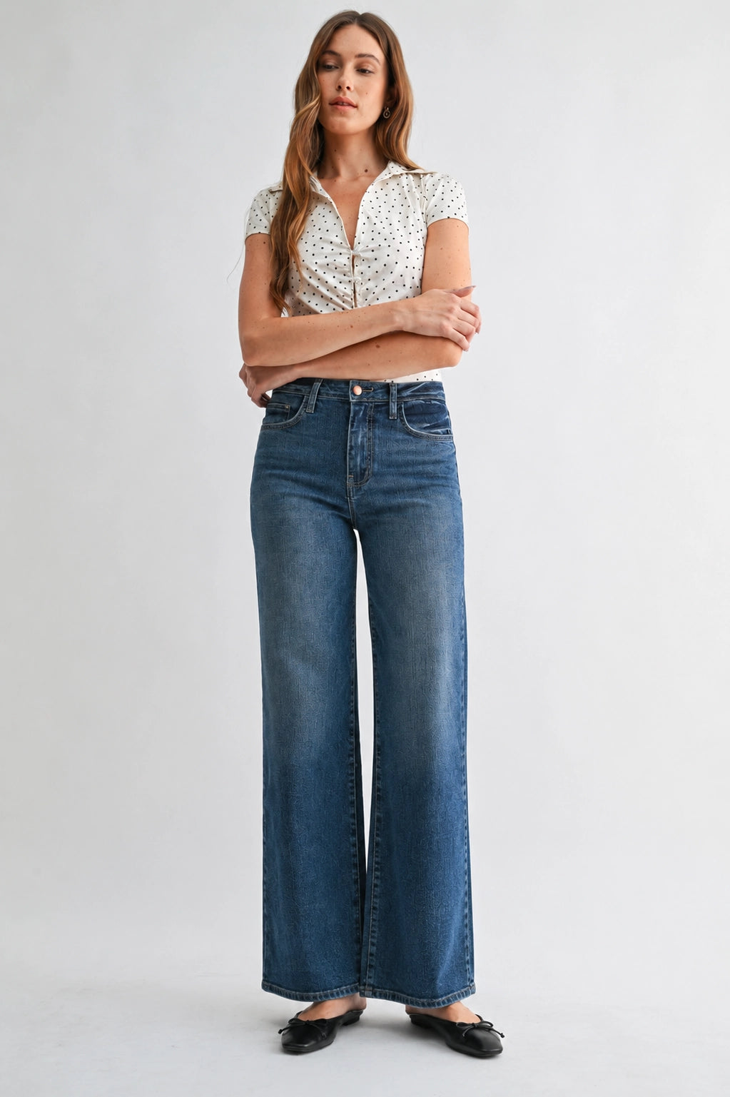 A woman modeling a pair of high-waisted, wide-leg blue denim jeans with a white patterned short-sleeve top and black flats, standing against a light gray studio background.