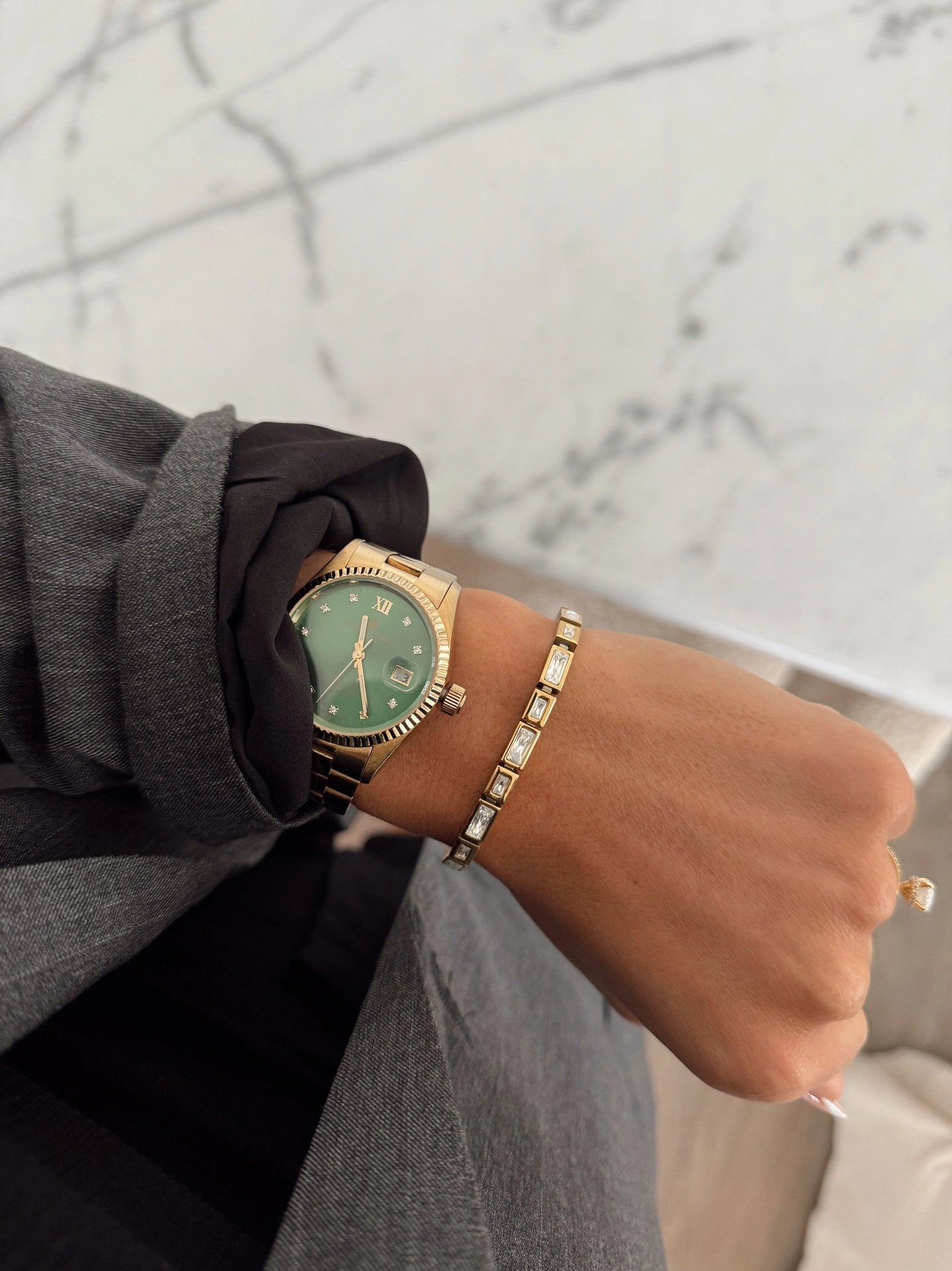 Hand wearing a gold watch with a green face and matching bracelet on a marble surface.