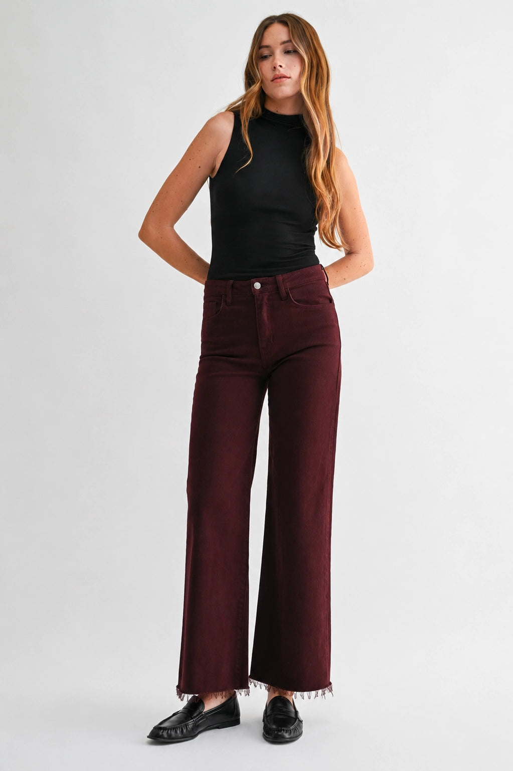 A woman modeling a pair of high-waisted, wide-leg maroon jeans with frayed hems, paired with a black sleeveless top and black loafers, against a light gray studio background.