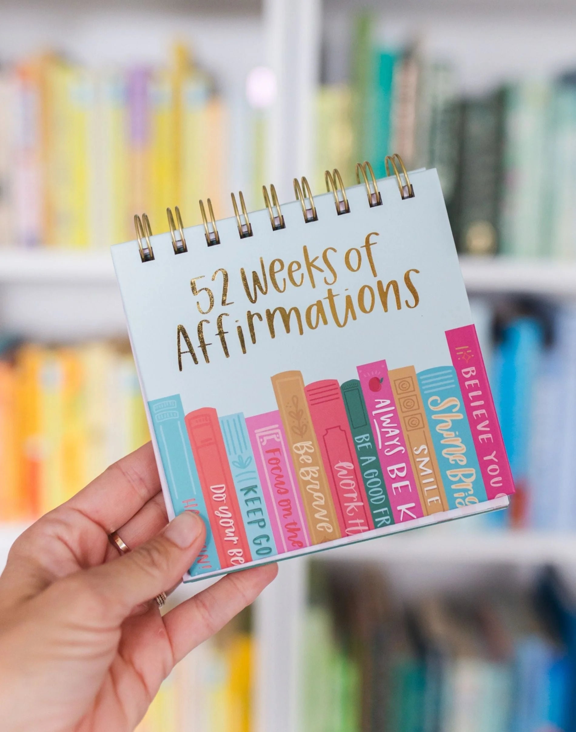 Hand holding a spiral-bound book titled '52 Weeks of Affirmations' with colorful text on a blurred library background.