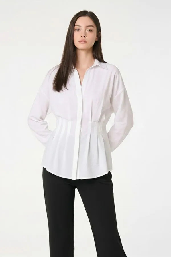 A woman modeling a white long-sleeve button-up blouse with a gathered waist, paired with black pants, against a plain white studio background.