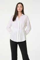 A woman modeling a white long-sleeve button-up blouse with a gathered waist, paired with black pants, against a plain white studio background.
