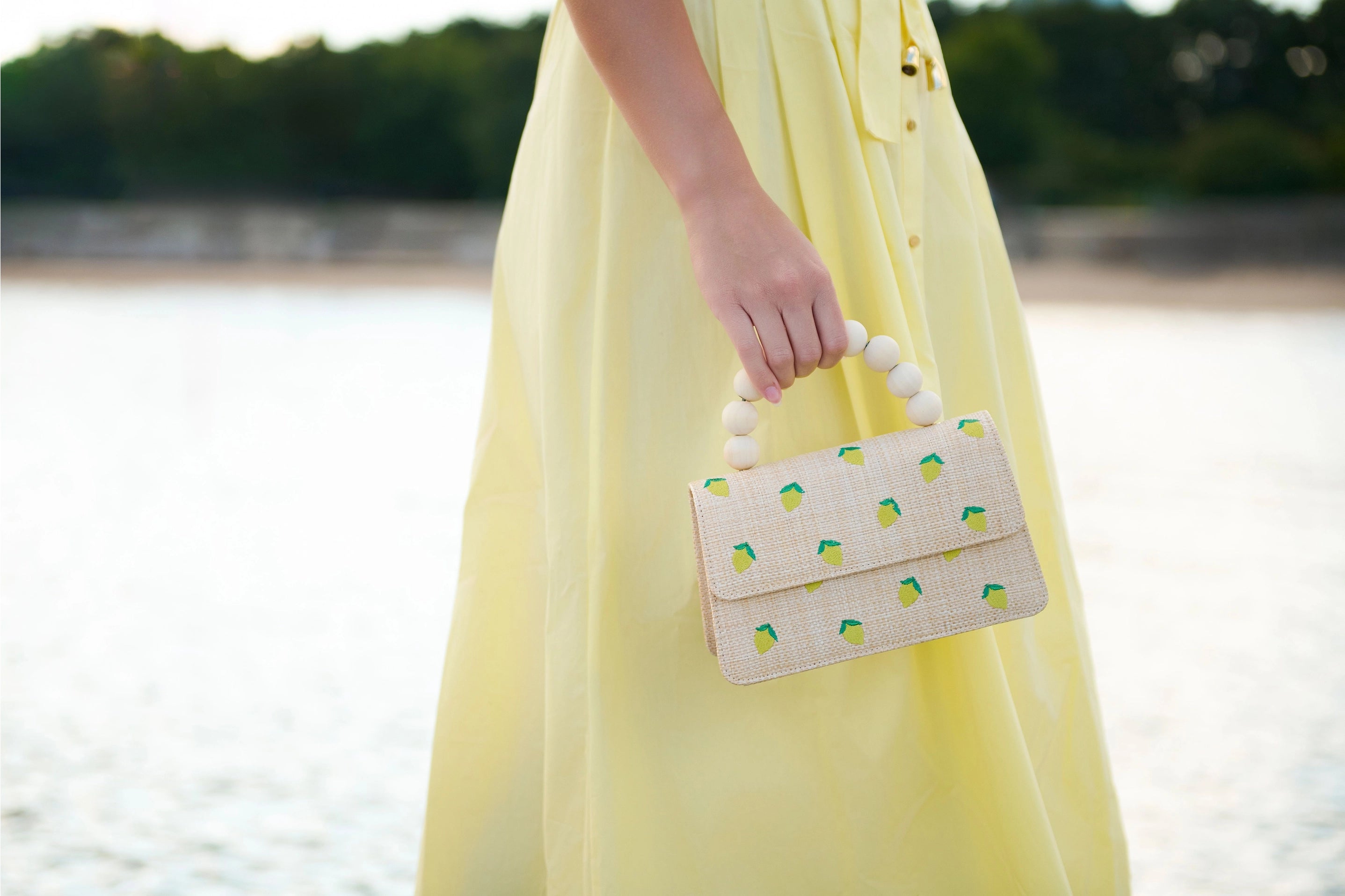 Person wearing a yellow dress holding a LEMON-patterned handbag by a body of water.