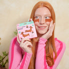 A woman with long red hair applies or wears brown paw-shaped eye patches while holding a pink package labeled 'paws' and 'spotted'. She is wearing a pink long-sleeve top against a beige textured wall.