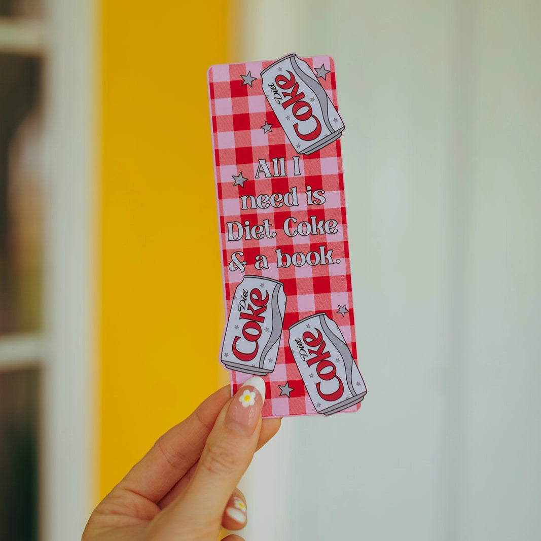 Hand holding a red and white checkered bookmark with Diet Coke and book text against a blurred background