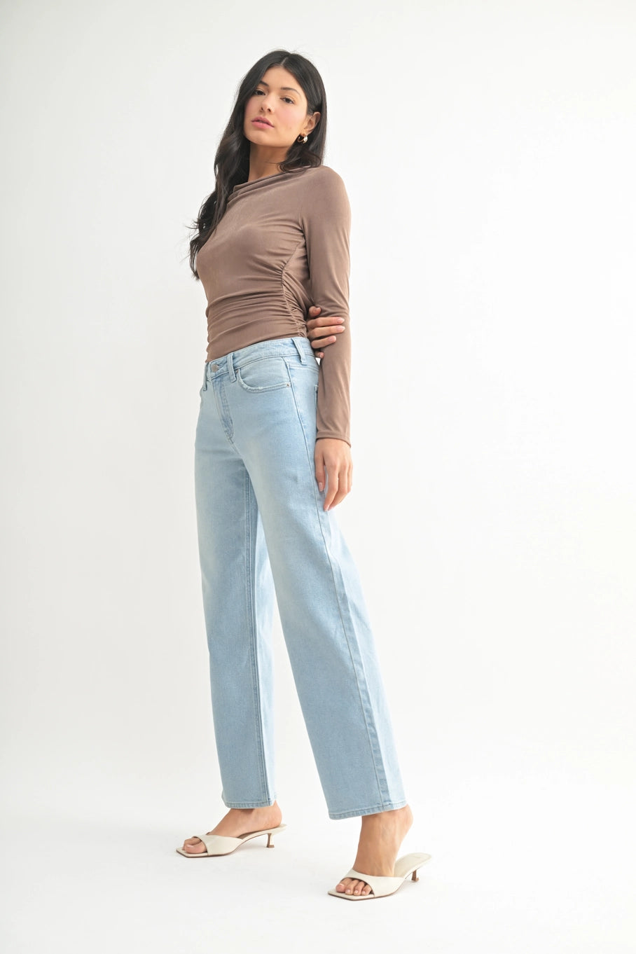 A woman modeling a brown long-sleeve top and light-wash blue straight-leg jeans with white heeled sandals, standing against a plain white studio background.