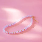 A delicate beaded necklace with pale pink translucent beads separated by small gold spacer rings, displayed on a soft pink background with artistic shadows.