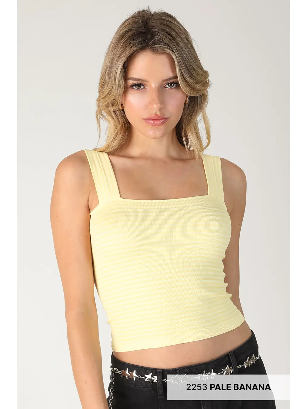 A woman modeling a pale yellow ribbed crop top with a square neckline and wide straps, paired with black studded jeans against a light gray studio background.