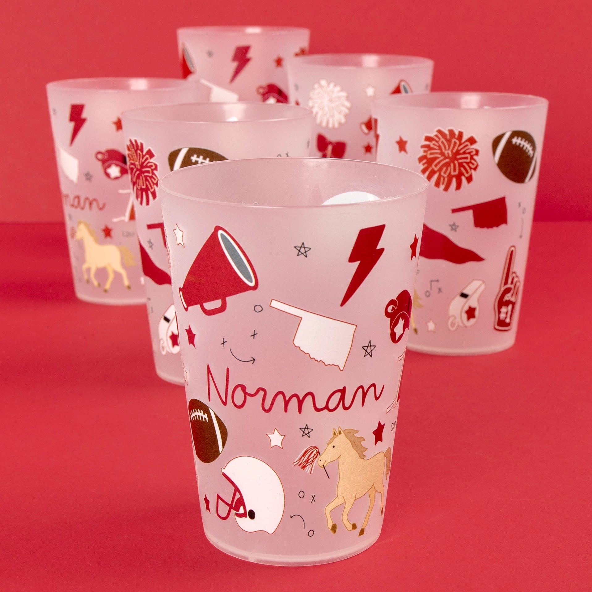 A set of frosted translucent plastic tumblers with red and brown sports-themed illustrations, including football, horses, stars, and the name 'Norman' on the front cup, displayed on a solid red background.