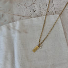A gold chain necklace with a small rectangular engraved gold tag pendant and a tiny clear stone charm, laid on a textured off-white linen fabric.