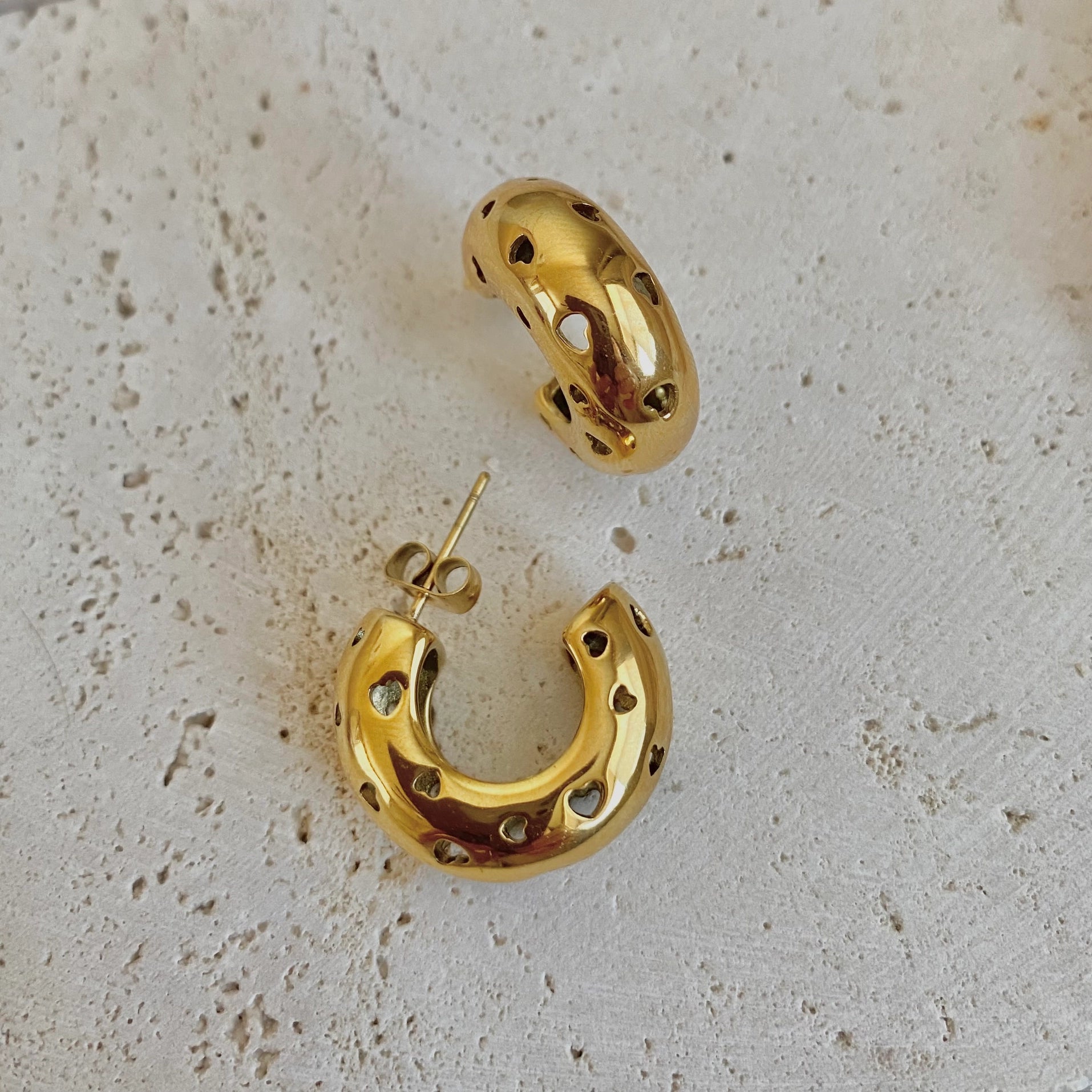 Gold hoop earrings with a textured, perforated design, displayed on a light beige stone surface.