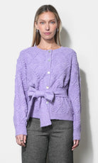 A woman modeling a lavender textured knit cardigan with a button-front closure and a matching tie belt at the waist, paired with gray trousers, against a plain light background.