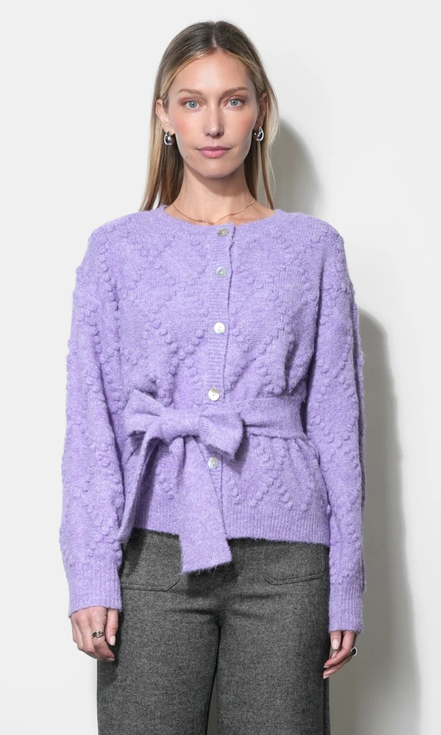 A woman modeling a lavender textured knit cardigan with a button-front closure and a matching tie belt at the waist, paired with gray trousers, against a plain light background.