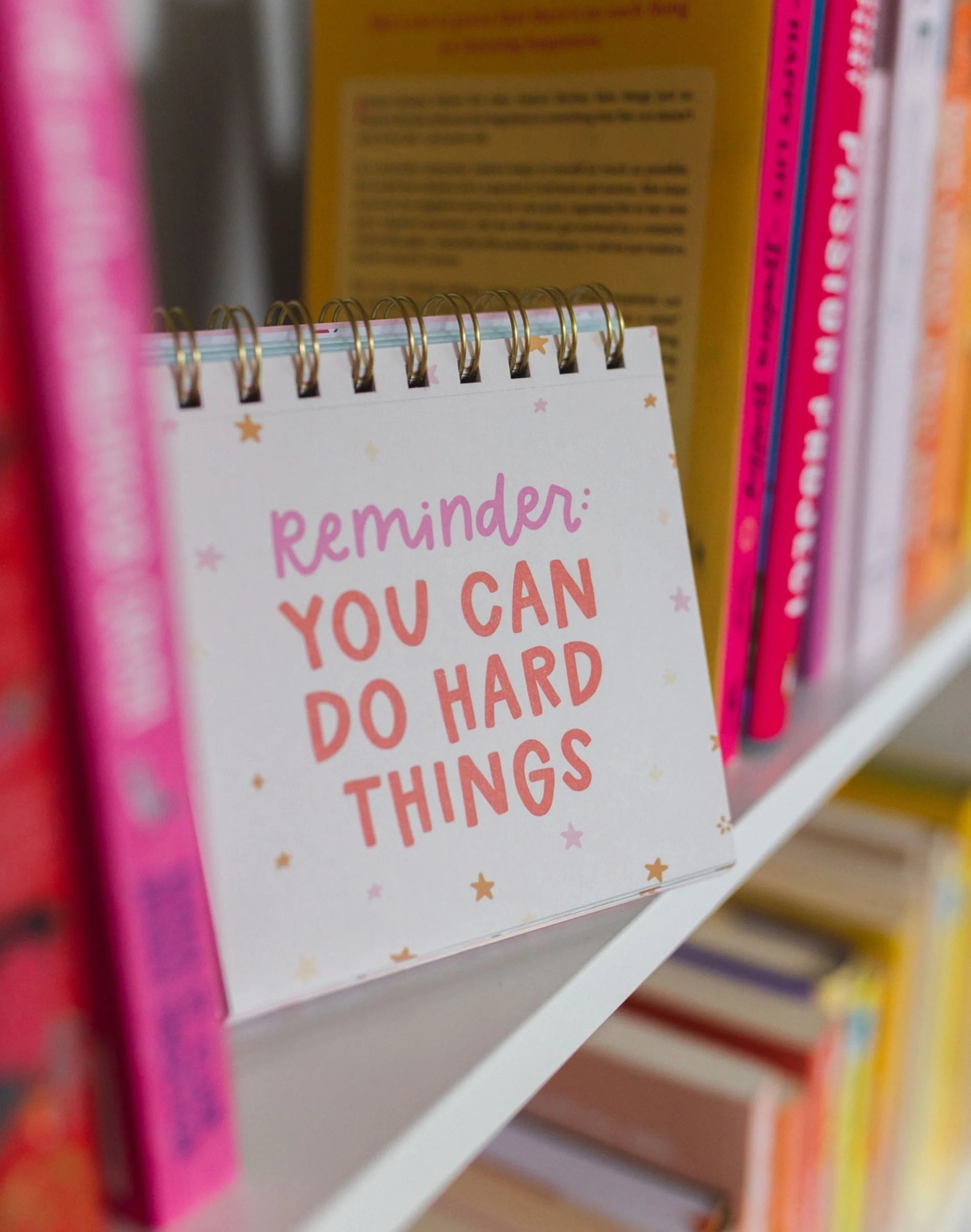 Notebook with 'Reminder: You can do hard things' text on a shelf with books.