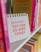 Notebook with 'Reminder: You can do hard things' text on a shelf with books.