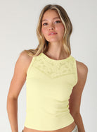 A woman modeling a pale yellow ribbed sleeveless top with a lace-like yoke detail, shown against a light gray studio background. 