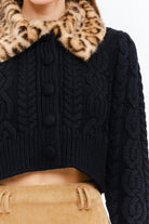 A close-up of a person wearing a black cable-knit cropped sweater with a leopard-print faux-fur collar and dark buttons, paired with a tan skirt.
