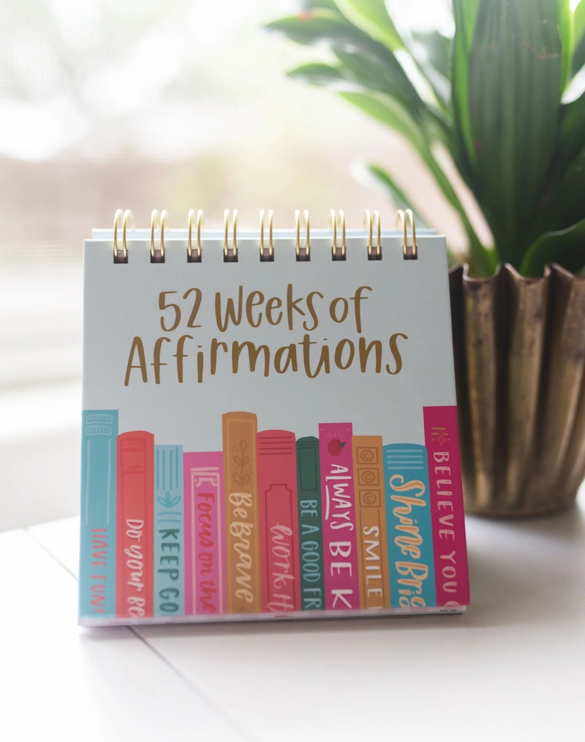 Small spiral-bound book titled '52 Weeks of Affirmations' on a surface with a plant in the background.