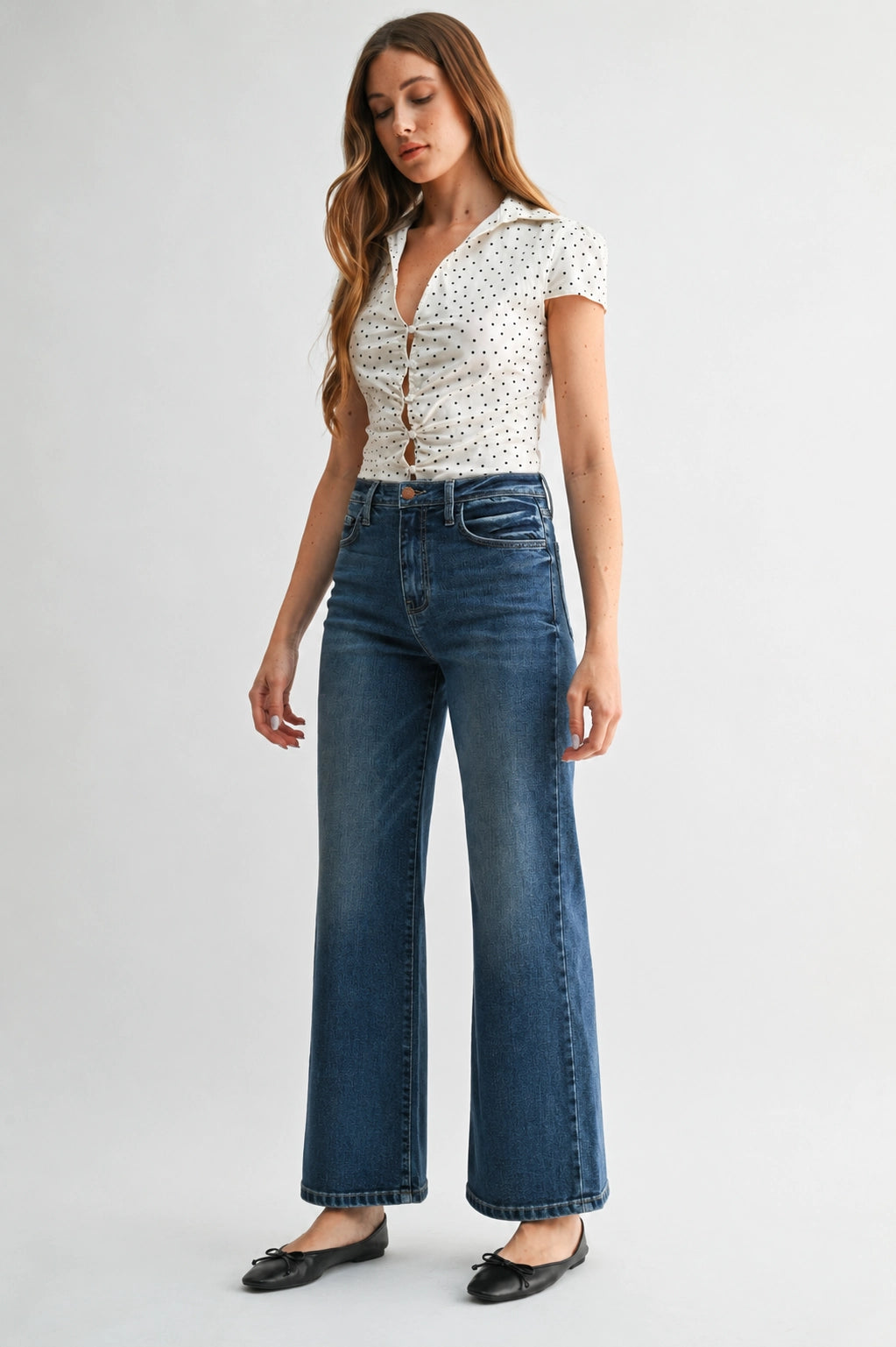 A woman modeling a pair of high-waisted, wide-leg blue jeans with a white short-sleeve button-up blouse featuring a black polka-dot pattern, paired with black flat shoes, against a light gray studio background.