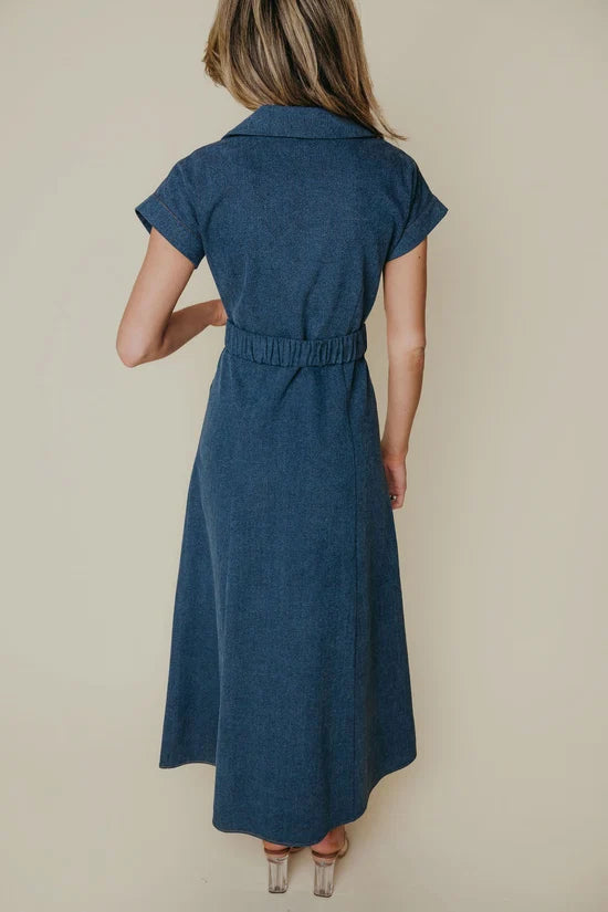 A woman viewed from the back wearing a short-sleeve blue midi dress with a collared neckline and a gathered elastic waist, standing against a plain beige studio background.