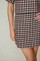 A close-up of a model wearing a short-sleeve mini dress with a black, white, and pink houndstooth pattern against a light beige background.