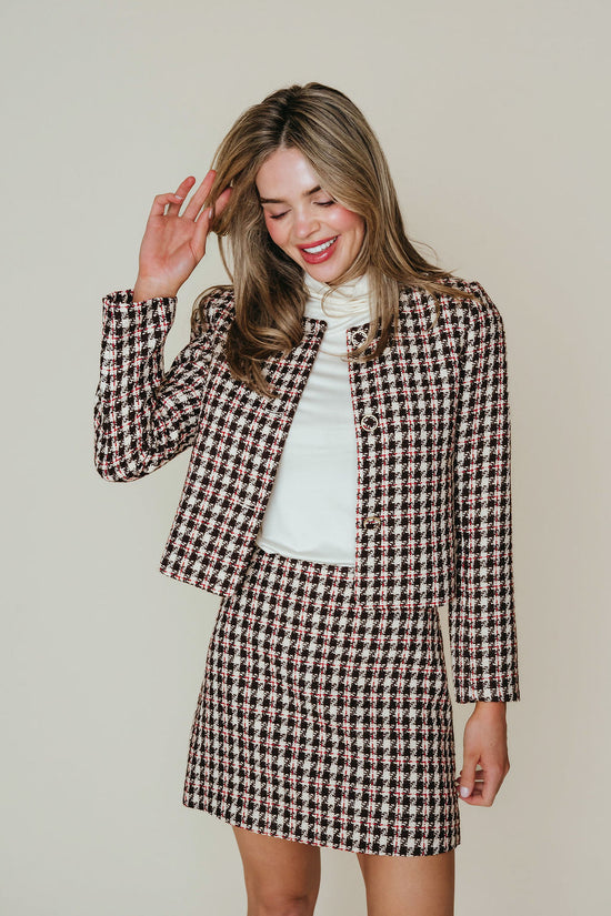 A woman modeling a matching houndstooth tweed jacket and mini skirt set over a white turtleneck, posing against a solid light beige background.