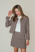 A woman modeling a matching houndstooth tweed jacket and mini skirt set over a white turtleneck, posing against a solid light beige background.